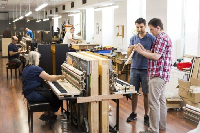 Diverse Expertinnen und Experten arbeiten in den Räumlichkeiten der Klaviermanufaktur an verschiedenen Klavieren